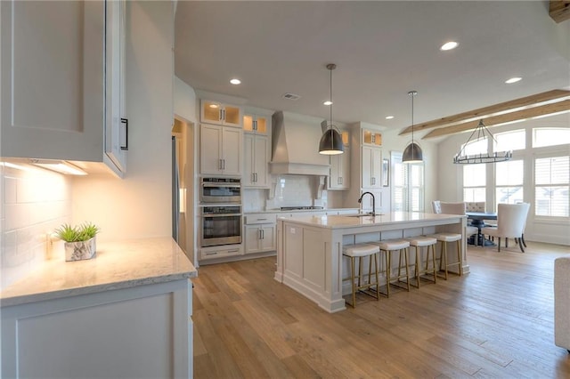 12605 W 169th St, Overland Park, KS 66221 - kitchen featuring custom range hood, tasteful backsplash, light wood-style flooring, a center island with sink, and white cabinets