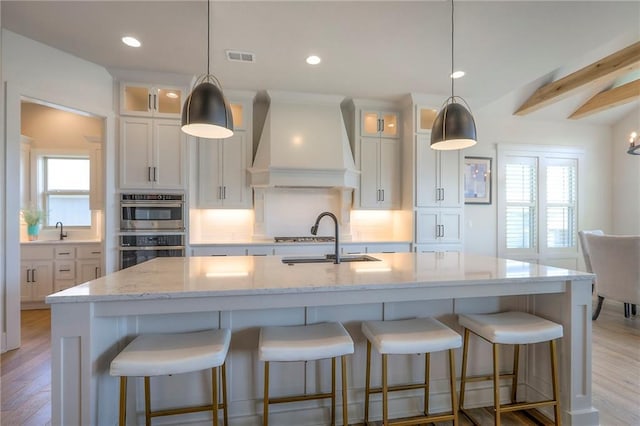 12605 W 169th St, Overland Park, KS 66221 - kitchen featuring custom exhaust hood, stainless steel double oven, an island with sink, white cabinets, and glass insert cabinets