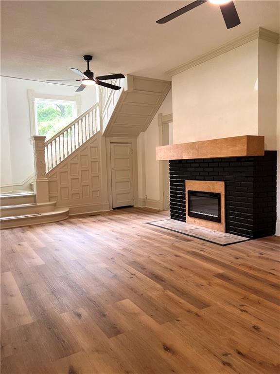 112 N High St, Butler, MO 64730 - unfurnished living room featuring ceiling fan, wood finished floors, a fireplace with flush hearth, ornamental molding, and a decorative wall