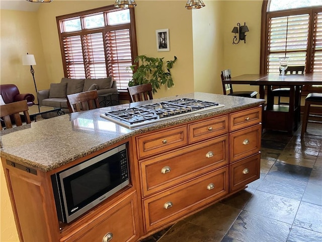 207 W 66th St, Kansas City, MO 64113 - kitchen with appliances with stainless steel finishes, brown cabinetry, stone tile flooring, a center island, and pendant lighting