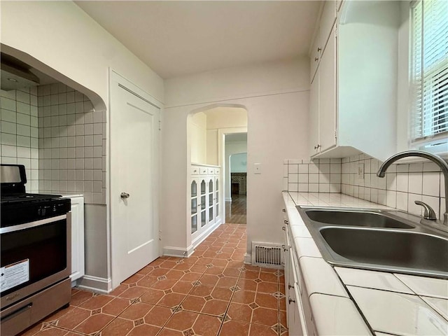 2416 E 70th St, Kansas City, MO 64132 - kitchen featuring stainless steel range with gas stovetop, tile countertops, white cabinets, arched walkways, and decorative backsplash