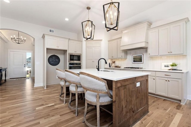 15323 W 171st Ter, Olathe, KS 66062 - kitchen with a chandelier, custom range hood, stainless steel microwave, an island with sink, and light wood-style flooring