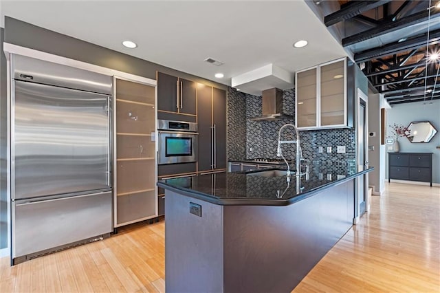 1303 W 47th St, Kansas City, MO 64112 - kitchen with stainless steel appliances, wall chimney exhaust hood, light wood-type flooring, dark stone counters, and tasteful backsplash