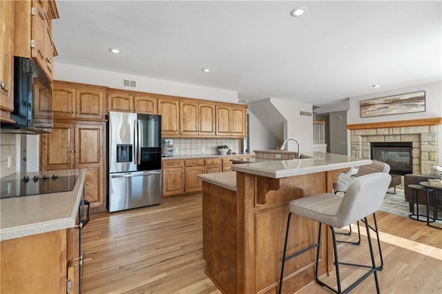 5701 W 146th St, Overland Park, KS 66223 - kitchen featuring black appliances, light wood-style flooring, a stone fireplace, a kitchen breakfast bar, and brown cabinetry