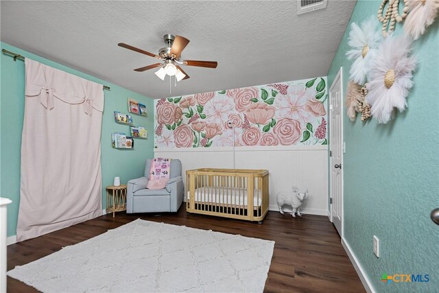unfurnished bedroom with a crib, a ceiling fan, a textured ceiling, wood finished floors, and wainscoting