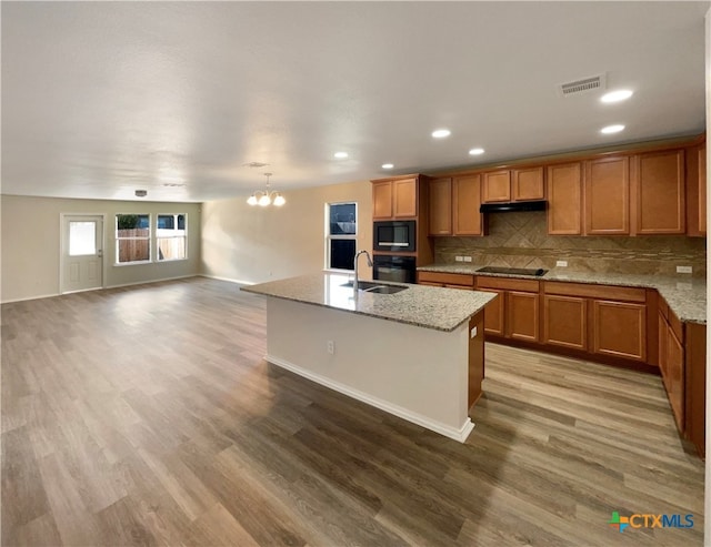 kitchen with black appliances, wood-type flooring, light stone counters, and a center island with sink