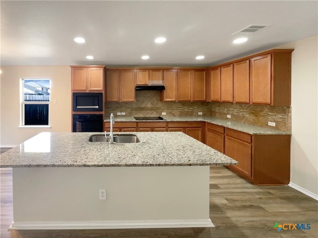 kitchen featuring black appliances, a center island with sink, light stone countertops, sink, and light hardwood / wood-style floors