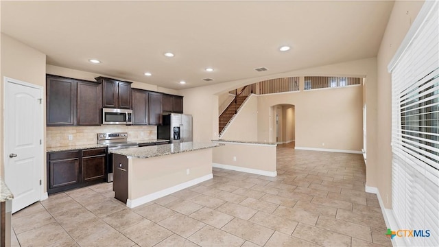 kitchen with decorative backsplash, appliances with stainless steel finishes, light stone counters, dark brown cabinets, and a kitchen island