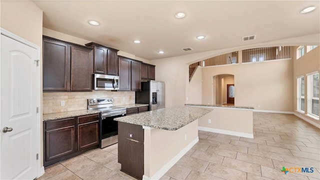 kitchen featuring dark brown cabinetry, light stone countertops, a center island, decorative backsplash, and appliances with stainless steel finishes