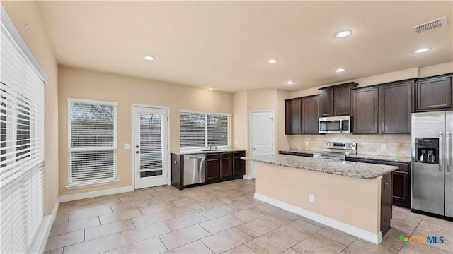 kitchen with appliances with stainless steel finishes, tasteful backsplash, dark brown cabinets, a kitchen island, and light stone counters