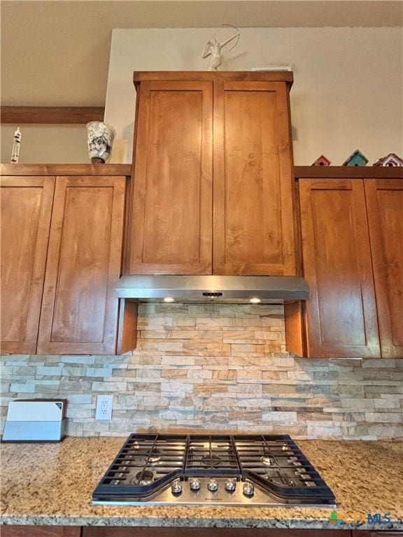 174 Riley Ln, Port Oconnor, TX 77982 - kitchen with stainless steel gas stovetop, brown cabinetry, backsplash, under cabinet range hood, and light stone counters