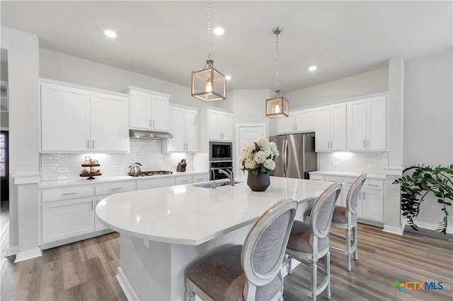 kitchen with a kitchen island with sink, under cabinet range hood, a sink, appliances with stainless steel finishes, and light countertops