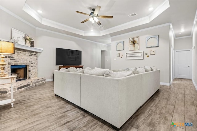 living room featuring a brick fireplace, crown molding, a raised ceiling, and ceiling fan