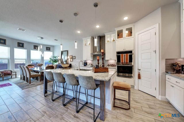 kitchen featuring double oven, sink, white cabinets, hanging light fixtures, and a center island with sink