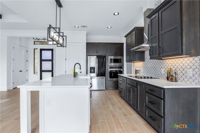 kitchen featuring backsplash, an island with sink, appliances with stainless steel finishes, decorative light fixtures, and light hardwood / wood-style floors