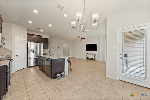 kitchen with stainless steel appliances, light stone counters, dark brown cabinetry, a kitchen island with sink, and decorative light fixtures