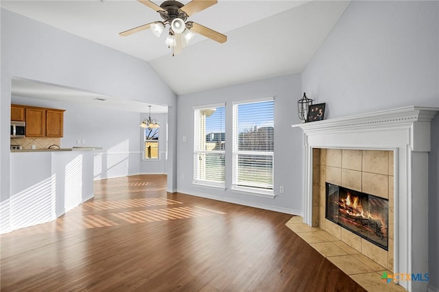 unfurnished living room featuring ceiling fan with notable chandelier, vaulted ceiling, a tile fireplace, and light wood-type flooring