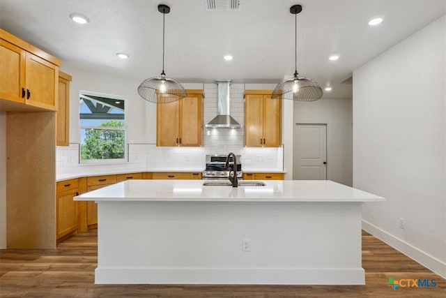 kitchen with hanging light fixtures, wall chimney exhaust hood, a kitchen island with sink, and hardwood / wood-style floors