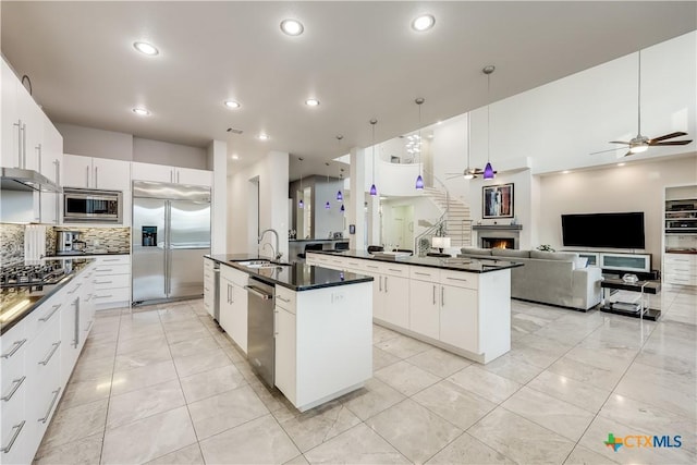 kitchen with a kitchen island with sink, sink, hanging light fixtures, built in appliances, and white cabinetry