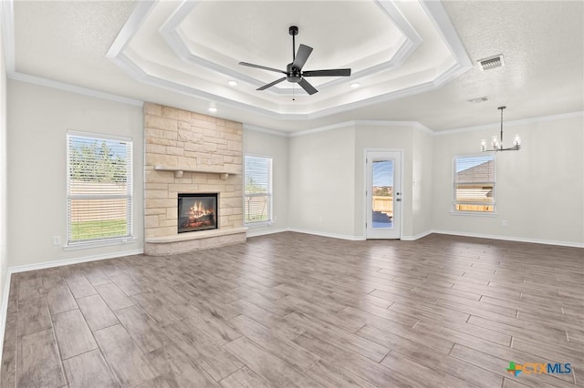 unfurnished living room with a raised ceiling, a stone fireplace, crown molding, and ceiling fan with notable chandelier