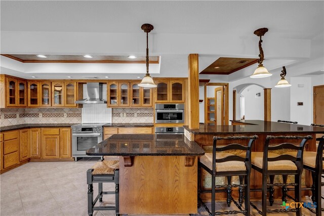 kitchen with a kitchen bar, stainless steel appliances, tasteful backsplash, wall chimney range hood, and pendant lighting