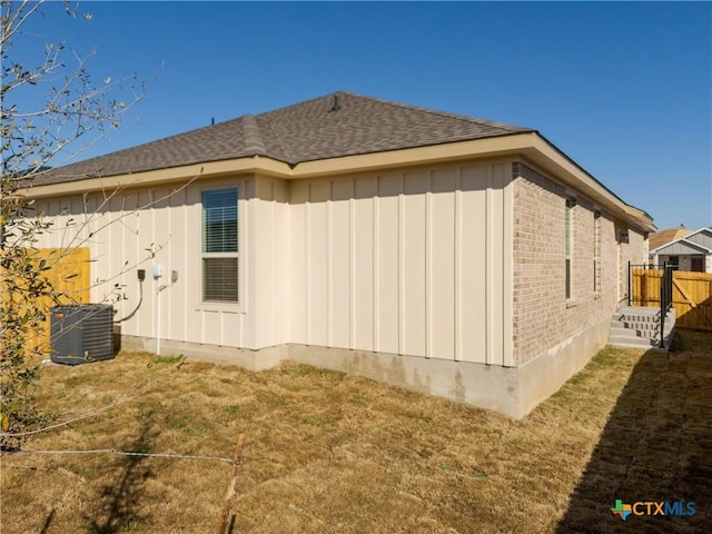 view of side of property with board and batten siding, a shingled roof, fence, central AC, and a yard