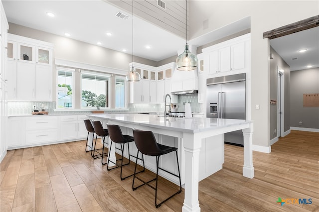 kitchen with built in fridge, white cabinetry, light wood-type flooring, and an island with sink