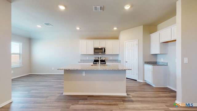 kitchen featuring a center island with sink, appliances with stainless steel finishes, light stone countertops, white cabinets, and light hardwood / wood-style flooring