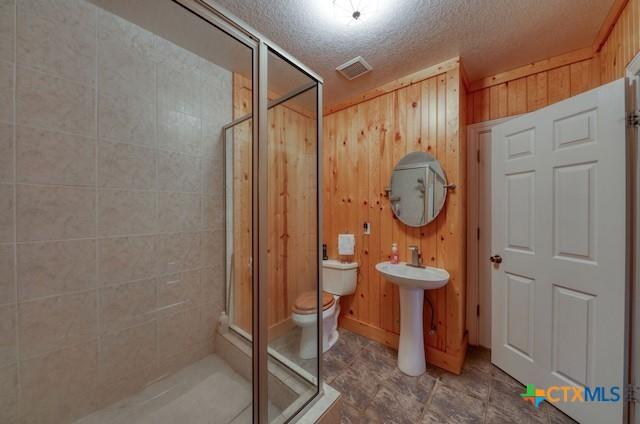 bathroom with a textured ceiling, a shower with door, wooden walls, sink, and toilet