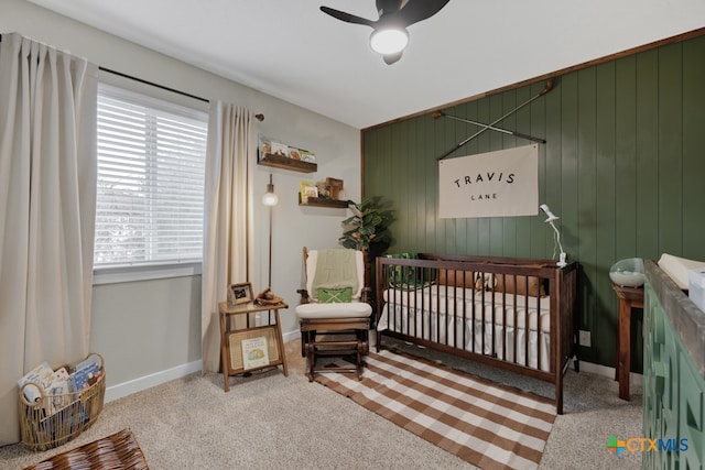 bedroom featuring a crib, wooden walls, ceiling fan, and carpet flooring