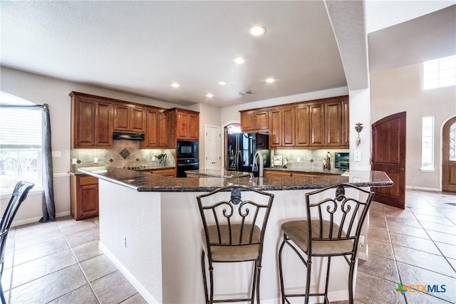 kitchen with a breakfast bar, black appliances, a center island with sink, dark stone counters, and backsplash