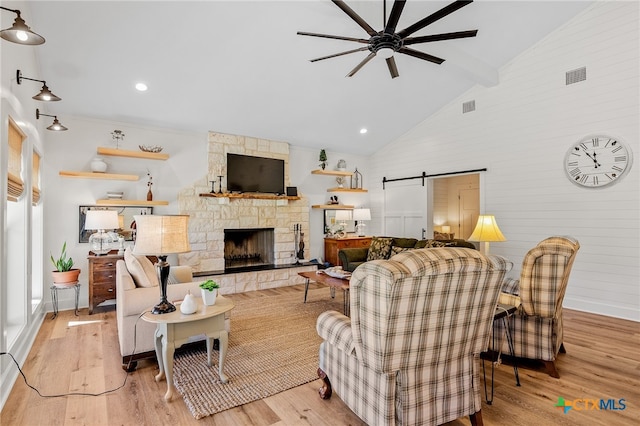 living room with ceiling fan, a barn door, high vaulted ceiling, light hardwood / wood-style floors, and a stone fireplace