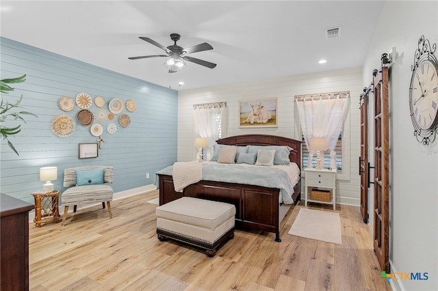 bedroom with a barn door, light hardwood / wood-style floors, ceiling fan, and wood walls