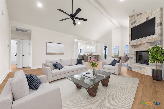 living room with ceiling fan, beam ceiling, high vaulted ceiling, light hardwood / wood-style flooring, and a stone fireplace