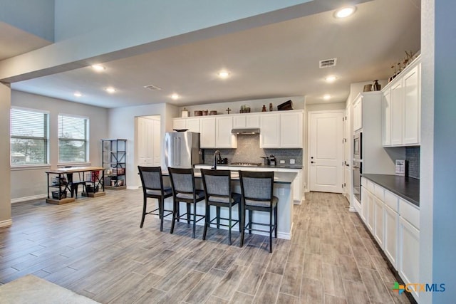 kitchen with stainless steel appliances, light hardwood / wood-style floors, white cabinets, a center island with sink, and decorative backsplash