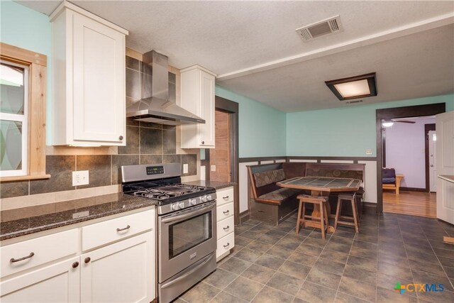 kitchen with backsplash, wall chimney range hood, stainless steel gas stove, dark stone countertops, and white cabinetry