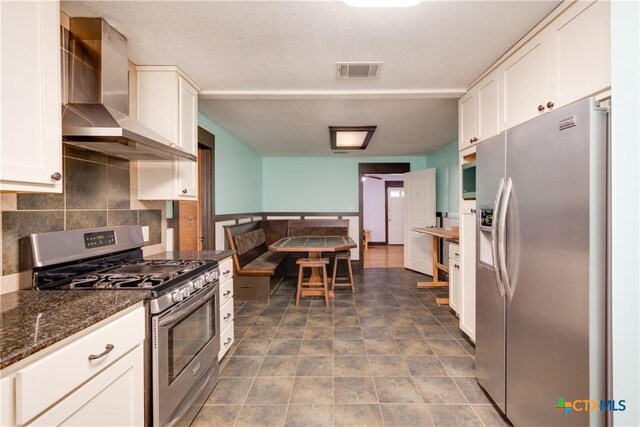 kitchen featuring appliances with stainless steel finishes, tasteful backsplash, wall chimney range hood, dark stone countertops, and white cabinets