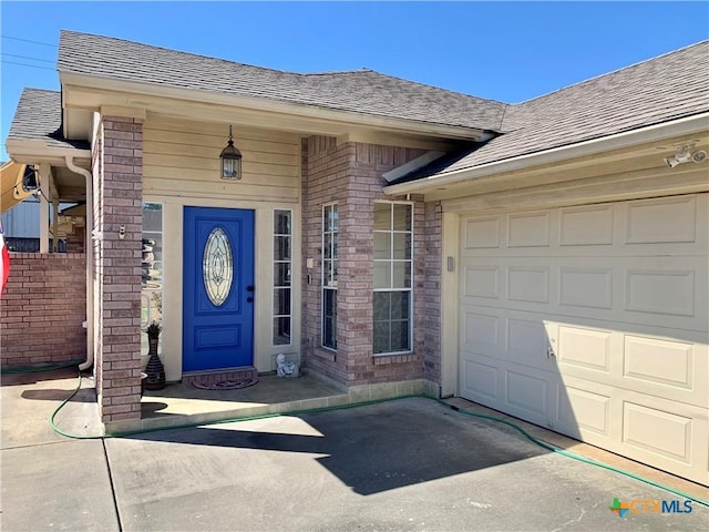 doorway to property featuring a garage, driveway, a shingled roof, and brick siding