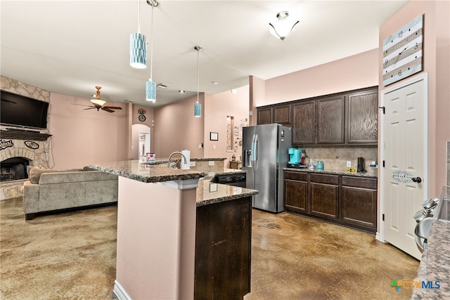 kitchen featuring dark brown cabinetry, hanging light fixtures, stainless steel fridge, light stone countertops, and a kitchen island with sink