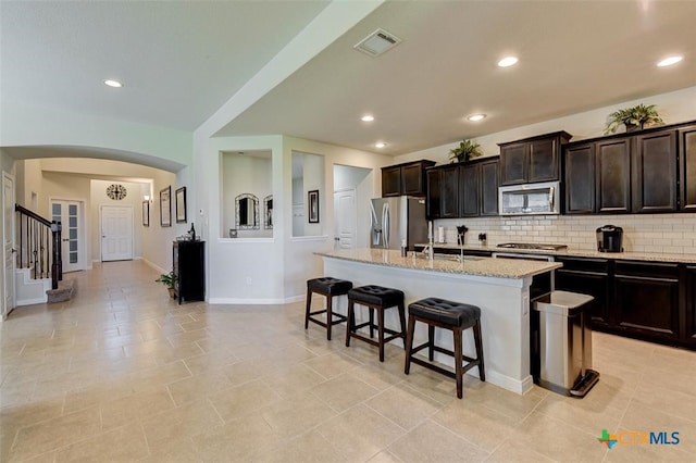 kitchen featuring decorative backsplash, a kitchen island with sink, a breakfast bar, light stone countertops, and appliances with stainless steel finishes