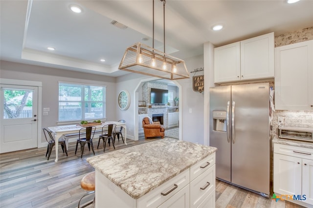 kitchen featuring pendant lighting, stainless steel refrigerator with ice dispenser, a large fireplace, white cabinets, and a raised ceiling