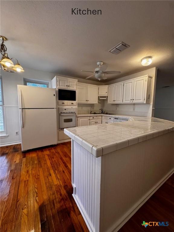 kitchen with tile countertops, dark wood-type flooring, white cabinets, white appliances, and a peninsula