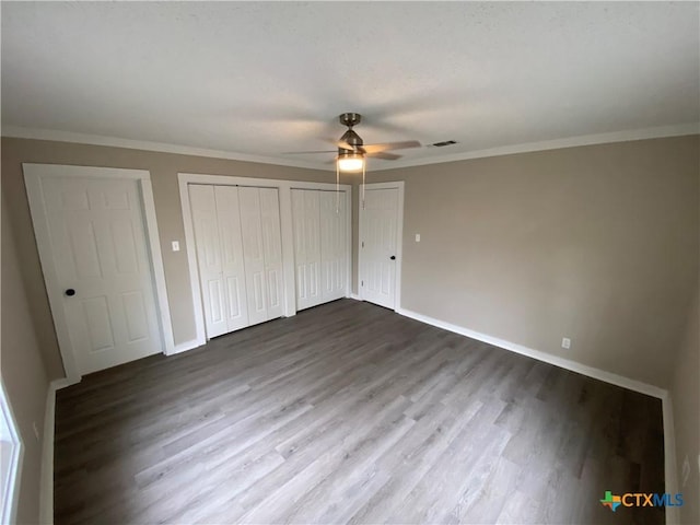 unfurnished bedroom with crown molding, dark wood finished floors, two closets, a ceiling fan, and baseboards