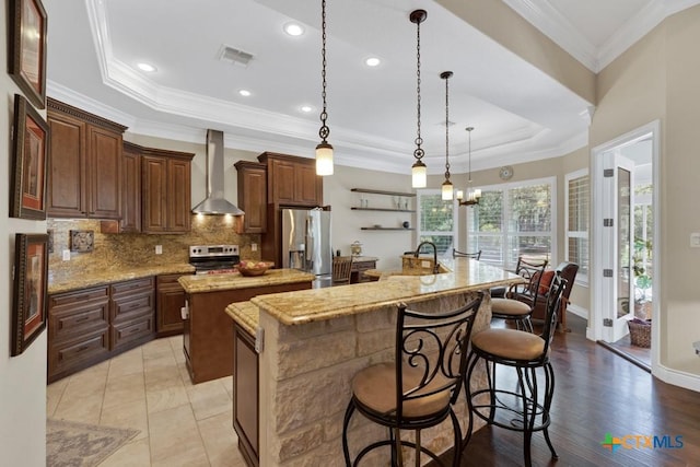 kitchen with appliances with stainless steel finishes, a raised ceiling, an island with sink, wall chimney exhaust hood, and visible vents