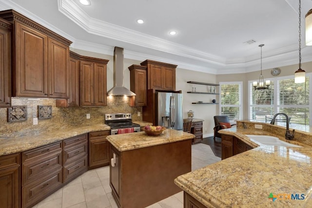 kitchen featuring an island with sink, wall chimney range hood, a tray ceiling, a sink, and stainless steel appliances
