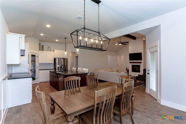 dining room with dark wood-type flooring, ceiling fan with notable chandelier, vaulted ceiling with beams, and a fireplace