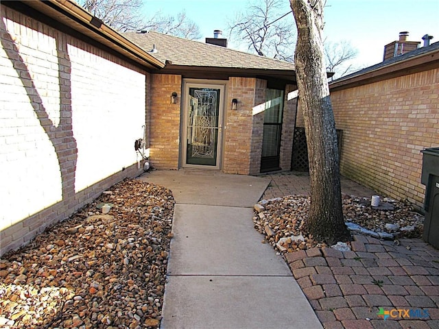 entrance to property with roof with shingles, brick siding, a chimney, and a patio area