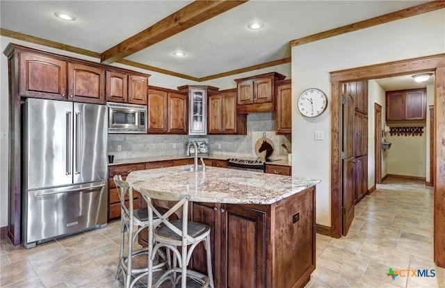 kitchen featuring beamed ceiling, light stone counters, appliances with stainless steel finishes, tasteful backsplash, and an island with sink