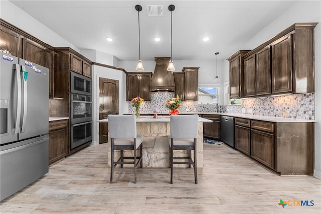 kitchen with light wood-type flooring, custom range hood, stainless steel appliances, decorative light fixtures, and a center island