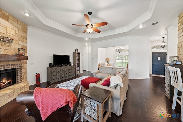 living room featuring ornamental molding, ceiling fan with notable chandelier, a tray ceiling, dark hardwood / wood-style floors, and a stone fireplace